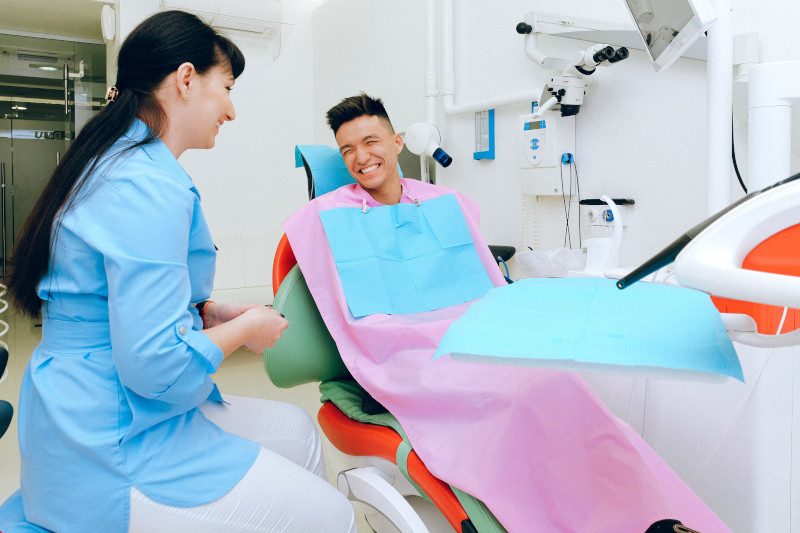 A young man sitting in a dentist’s examination chair, smiling, while a dentist sits on a stool next to him, also smiling.