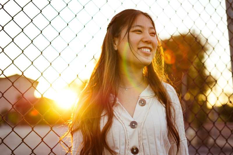 A woman standing against a fence, smiling during a sunset.